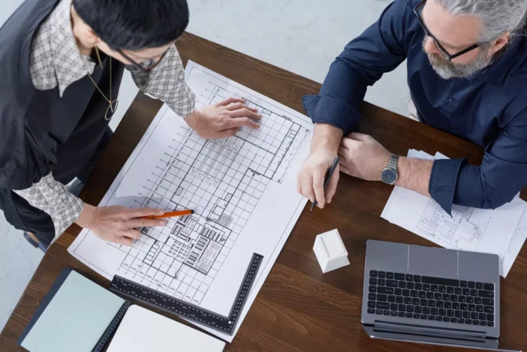 Two professionals discussing architectural blueprints on a wooden table, with a laptop, scale ruler, and a small building model, representing BIM Estimating Services.